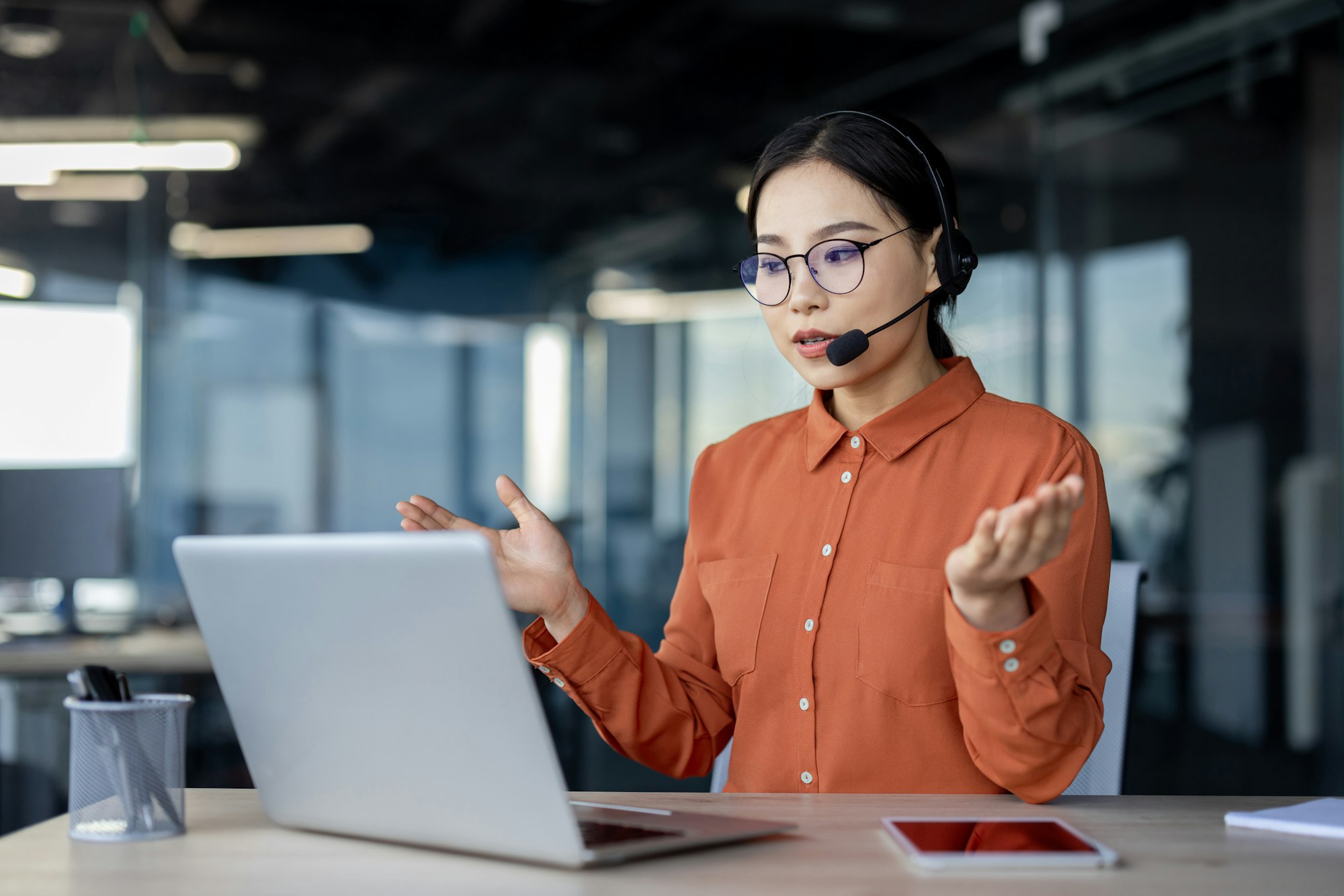 Serious asian woman with headset talking remotely using laptop for video call, help desk worker